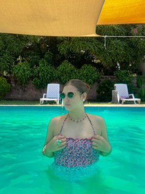 Renkli bir mayo giymiş genç bir kadın, parlak bir havuzda, sarı bir gölgelik ve yemyeşil bir arka planda, şakacı bir şekilde işaret ediyor. Yaz, yaşam tarzı ve tatil temalı projeler için mükemmel..