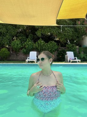 Renkli bir mayo giymiş genç bir kadın, parlak bir havuzda, sarı bir gölgelik ve yemyeşil bir arka planda, şakacı bir şekilde işaret ediyor. Yaz, yaşam tarzı ve tatil temalı projeler için mükemmel..