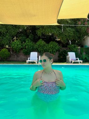 Renkli bir mayo giymiş genç bir kadın, parlak bir havuzda, sarı bir gölgelik ve yemyeşil bir arka planda, şakacı bir şekilde işaret ediyor. Yaz, yaşam tarzı ve tatil temalı projeler için mükemmel..