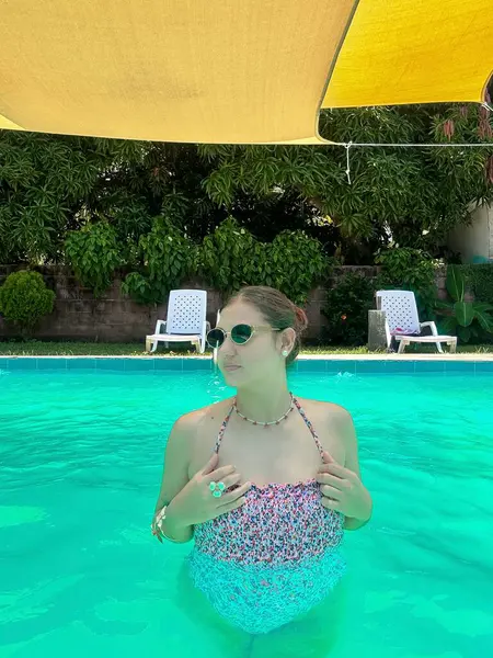 Renkli bir mayo giymiş genç bir kadın, parlak bir havuzda, sarı bir gölgelik ve yemyeşil bir arka planda, şakacı bir şekilde işaret ediyor. Yaz, yaşam tarzı ve tatil temalı projeler için mükemmel..