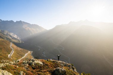 İsviçre 'nin İsviçre Alpleri' ndeki Furka Geçidi ve Grimselpass 'ın virajlı yollarına bakan uçurumun kenarında ellerini kaldırmış bir kadın silueti. Sabahın erken saatlerinde dağların tepelerinde.