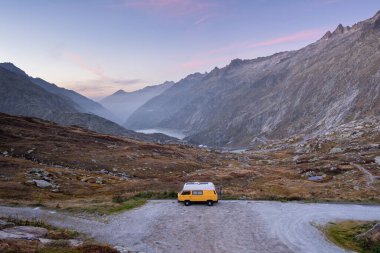 İsviçre 'nin Alp Dağları' ndaki Grimsel Geçidi 'ne park edilmiş klasik sarı bir karavan. Arka planda kayalık dağlar ve mavi gökyüzü.