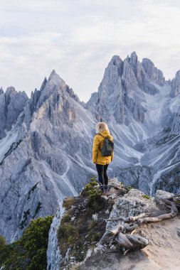 Dolomite Alpleri 'nde Tre Cime di Lavaredo Dağı yakınlarında yürüyüş yapan sarı yağmurluklu kadın. Sonbahar günü ve tepelerin üstündeki açık gökyüzü.