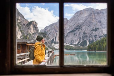 Woman in yellow raincoat enjoying the scenery at Lake Braies, Prags Dolomites in South Tyrol, Italy. View through the window over the boathouse and mountains. clipart