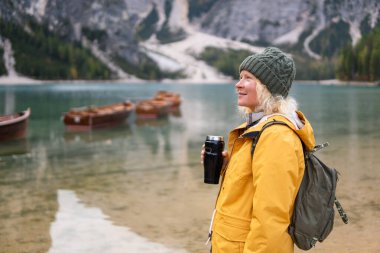 Sarı yağmurluklu kadın Braies Gölü 'nde termo bardak tutuyor Güney Tyrol, İtalya' da Prags Dolomitleri. Arka planda tahta kayıklar var..