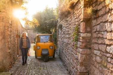 Young blonde woman walking the cobbled streets of a hilltop town in Tuscany with an old auto rickshaw on the background, Italy. clipart