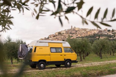 İtalya 'nın Umbria şehrinde arka planda tepe tepesi olan zeytin ağacı bahçesinden geçen klasik sarı bir karavan..