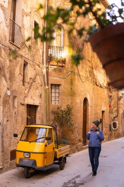 Genç sarışın kadın, Tuscany 'nin yamaçlı sokaklarında yürüyor. Arka planda eski bir fayton var, İtalya. Dikey yönelim
