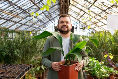 Serada çalışan genç bir adamın portresi. Kameraya bakıyor, gülümsüyor ve elinde büyük bir saksı tutuyor..