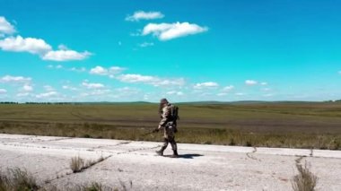 Yalnız ve yorgun bir asker mavi gökyüzüne karşı eski beton bir yolda yürüyor. 4K 'lık bir görüntü. Arka taraf görünümü.