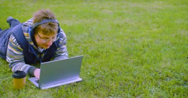 Kulaklık takan bir genç diz üstü bilgisayar monitörünün önünde çimlere uzanır. Ödevini yapar ya da sosyal ağlardan arkadaşlarıyla internetten iletişim kurar. 4k görüntü.