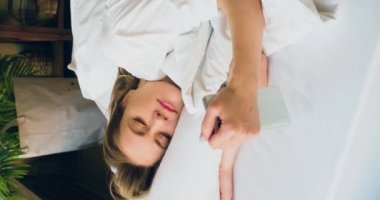 35 yaşında bir kadının uyandığı, akıllı telefonundaki mesajları kontrol ettiği ve tekrar uyuduğu yakın plan görüntüsü. Sosyal medya bağımlılığı kavramı. Dikey 4k görüntü.