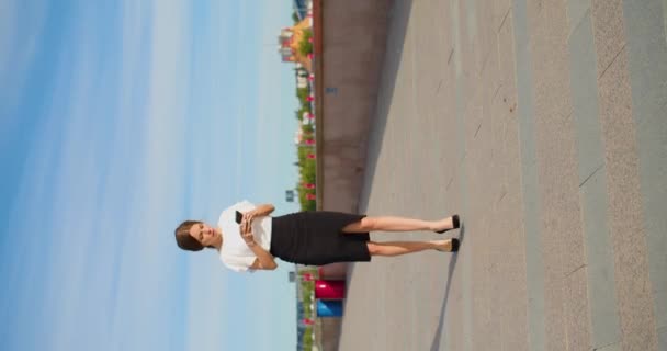 若い実業家が堤防に沿って歩き メッセージを受け取ります 素晴らしいニュース 彼女は喜んで面白いダンスを路上で右 縦4K映像 — ストック動画