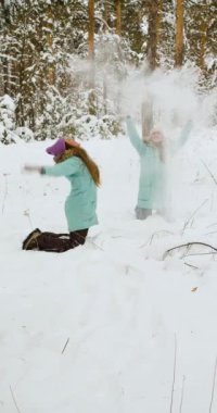 Karlı bir ormanda iki genç kadın eğleniyor. Onlar kış tatillerinde kar atar ve eğlenirler. Dikey video. Yavaş çekim 4k görüntü.
