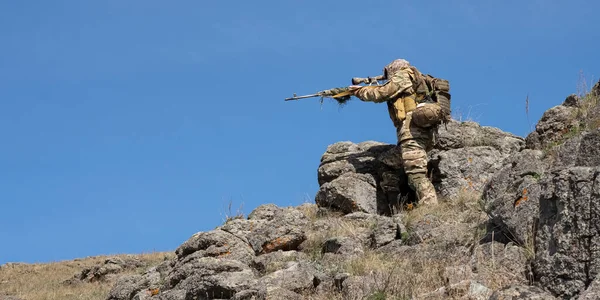 Ambush in the mountains - special forces professional sniper aiming at the enemy in the mountains. Concept of modern military operations and a special operation on enemy territory. Format photo 2x1.