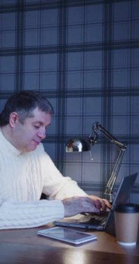 Orta yaşlı bir adam gece geç saatlerde bir bilgisayarın önünde çalışır. Bir elektrik lambasının ışığında metin yazdırıyor. Dikey 4k video.