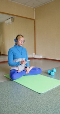 Kulaklık takan genç bir kadın müziği yatıştırmak için meditasyon yapıyor, akıllı telefonundaki bir internet uygulaması ile müziği açıyor ve meditasyona başlıyor. Dikey 4k görüntü.