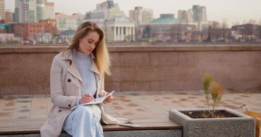 35 yaşında sarışın, açık havada çalışıyor. Akıllı telefonuyla e-postalarını kontrol ediyor ve not defterine not alıyor. 4k görüntü.