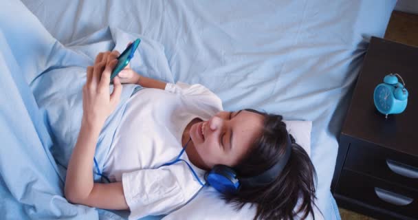 10岁的女孩拿着智能手机躺在床上 她正在听音乐或在醒来后玩游戏 现代技术概念 现代技术概念 慢动作4K镜头顶部视图 — 图库视频影像