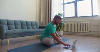İnternette yoga dersi alan orta yaşlı bir adamın fotoğrafı. Dizüstü bilgisayar monitörünün önünde yerde oturmuş meditasyon yapıyor. 4K görüntü.