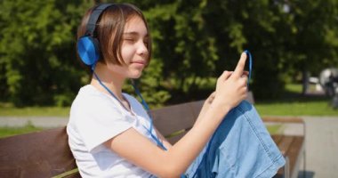 A teenage girl listens to music in headphones through an online smartphone application. Outdoor lifestyle close-up portrait. 4k footage.