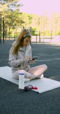 Young woman meditating after outdoor workout. She sits in headphones and listens to relaxing music. Vertical 4k footage.
