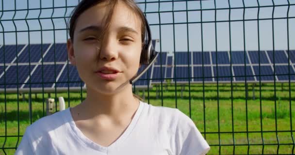 一名戴耳机的少女向学校报告了可持续能源生产的情况 她站在一个太阳能电池板农场附近 主持她的在线流媒体 4K镜头 — 图库视频影像