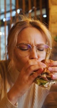 Close-up view of a young woman eating hamburger at a cafe. Slow motion vertical 4k footage.