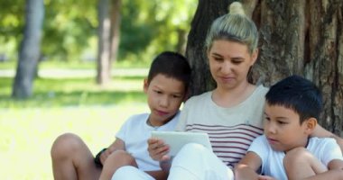 Close-up video of an Asian woman with two children watching online video while sitting under a tree in the park. A family watches a video through a tablet while sitting under the tree.