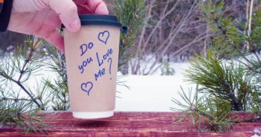 Male and female hands put paper cup with coffee on the wooden board in winter forest with inscriptions do you love me and yes. Love and relationships concept. Slow motion 4k footage.