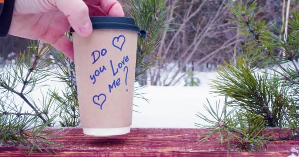 男性と女性の手は碑文と冬の森の中の木の板の上にコーヒーと紙コップを入れ あなたは私を愛していますか 愛と関係の概念 スローモーション4K映像 — ストック動画