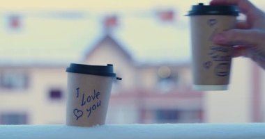 I love you and I love you too - male hand puts a glass of coffee in the snow with inscriptions. The concept of love and relationships. 4k slow motion.