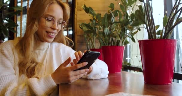 若い女性がカフェで注文を待っている間 スマートフォンアプリを介してオンラインで通信します — ストック動画