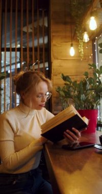 Young woman in reading book while sitting at a cafe. Slow motion vertical 4k footage.