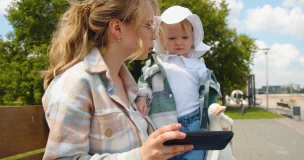 散歩中の子供を持つ若い母親 彼らはスマートフォンのオンラインアプリケーションを通じて子供のための楽しい教育ビデオを見ています 4K映像 — ストック動画