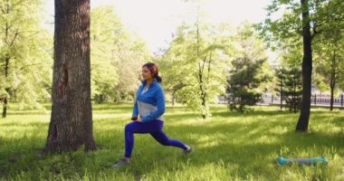 A 30-year-old woman is warming up in city park. She makes a lunge stretching and warming up the muscles of the legs. 4k footage.