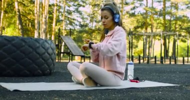 A young woman chooses music for meditation on her smart watch and starts meditating. Outdoor workout concept - meditation and mental health. 4k footage.