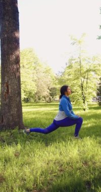 Vertical video of a 30-year-old woman is warming up in the park. She makes a lunge stretching and warming up the muscles of the legs. 4k footage.