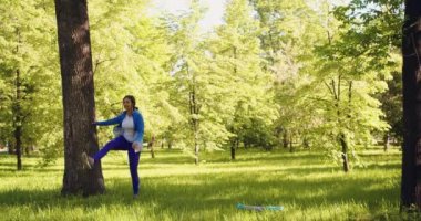 A 35-year-old woman warms up her muscles before a workout in a city park. She makes inertial swings with her foot. 4k footage.