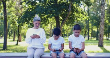 Asian family with smartphones - mother and two sons sit buried in smartphones outdoor. Smartphone addiction. 4k footage.