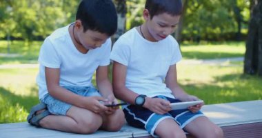 Close-up view of a two boys are playing on their smartphones while walking outdoors - they are sitting on a bench, looking at their smartphones. 4k footage.