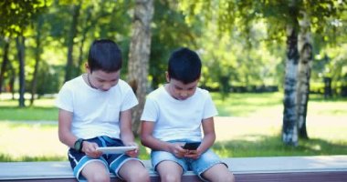 Two boys are playing on their smartphones while walking outdoors - they are sitting on a bench, looking at their smartphones. 4k footage.