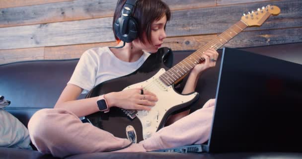 自宅で小さな女性ギタリスト ヘッドフォンの女の子はノートパソコンのモニターの前でエレキギターで家に座っている — ストック動画