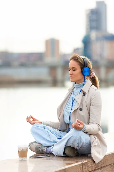 A young woman meditates on the embankment - she sitting in headphones and listening to relaxing music. Rest from office bustle and city noise. Vertical photo.