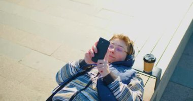 A teenage boy in headphones during online chat while lies on a bench whith smartphone. Close-up 4k footage.