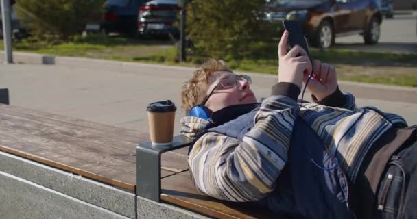 Kulaklık Takan Genç Bir Çocuk Bir Bankta Uzanır Akıllı Telefon — Stok video