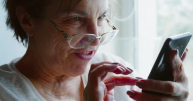 Close-up of an elderly woman in eyeglasses receives a funny message on her smartphone and laughs happily. Close-up 4k footage.