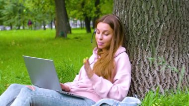 Smiling woman with laptop and headset having online conversation while sitting in city park - watching educational webinar on laptop. Slow motion footage.