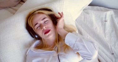 A woman listens to music through headphones while lying in bed. Modern technologies in the concept of everyday life. Slow motion 4k footage.