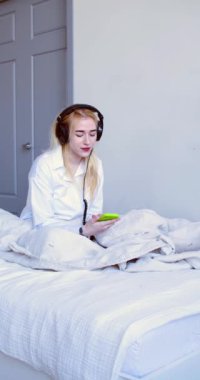 Vertical footage of a young woman woke up and listens to music through headphones with online smartphone app while sitting in bed. Modern technology in everyday life concept.
